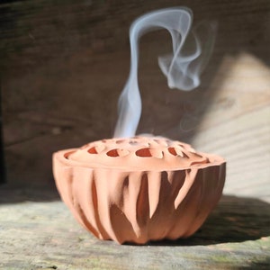 Puede incluir: Un quemador de incienso de terracota con forma de flor de loto con humo saliendo de la parte superior. El quemador tiene una superficie texturizada y está decorado con pequeños agujeros.