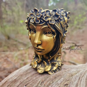 May include: A gold-colored planter shaped like a woman's face, adorned with a floral crown and leaf accents. The planter is set on a piece of wood, with a blurred forest background. The hashtag #tindymephate is visible.