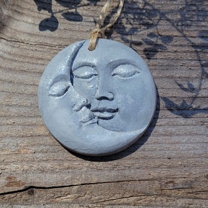 May include: A gray ceramic ornament shaped like a crescent moon with a face on the full side. The ornament is hanging from a twine string.
