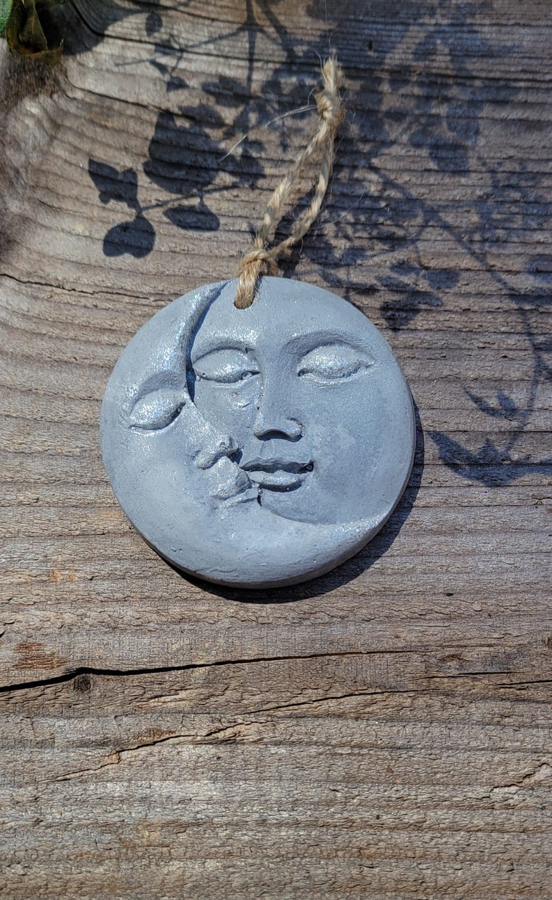 May include: A gray ceramic ornament shaped like a crescent moon with a face on the full side. The ornament is hanging from a twine string.