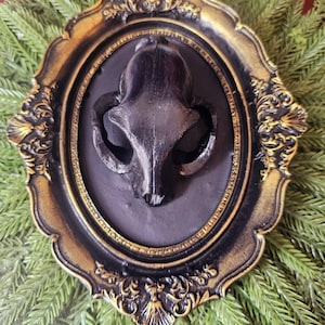 May include: A black skull sculpture is set within an ornate, decorative frame. The frame is black with gold detailing, and the skull is mounted on a dark background. This is likely a gothic or Halloween-themed home decor item.