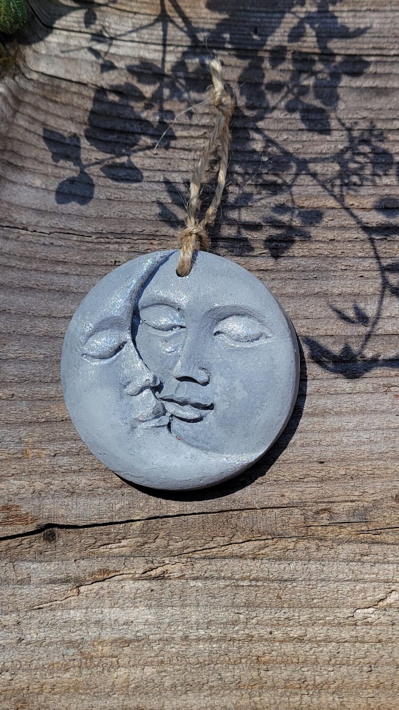 May include: A gray ceramic ornament shaped like a crescent moon with two faces, one on each side. The ornament is hanging from a piece of twine.