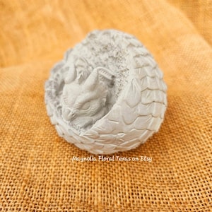Pode incluir: Pequena estatueta de dragão cinza claro enrolada em uma bola. O dragão está aninhado dentro de uma concha texturizada e escamosa. O texto "Magnolia Floral Texas on Etsy" é visível na superfície.