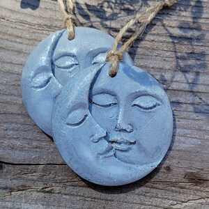 May include: Two gray painted ceramic moon ornaments with faces and a crescent moon design. The ornaments are hanging from twine.