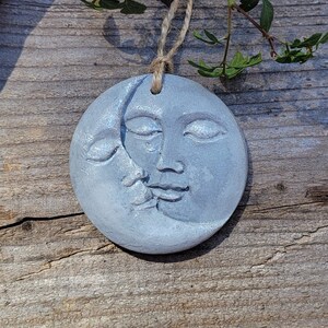 May include: A gray ceramic ornament shaped like a crescent moon with a face on it. The moon is hanging from a twine string.