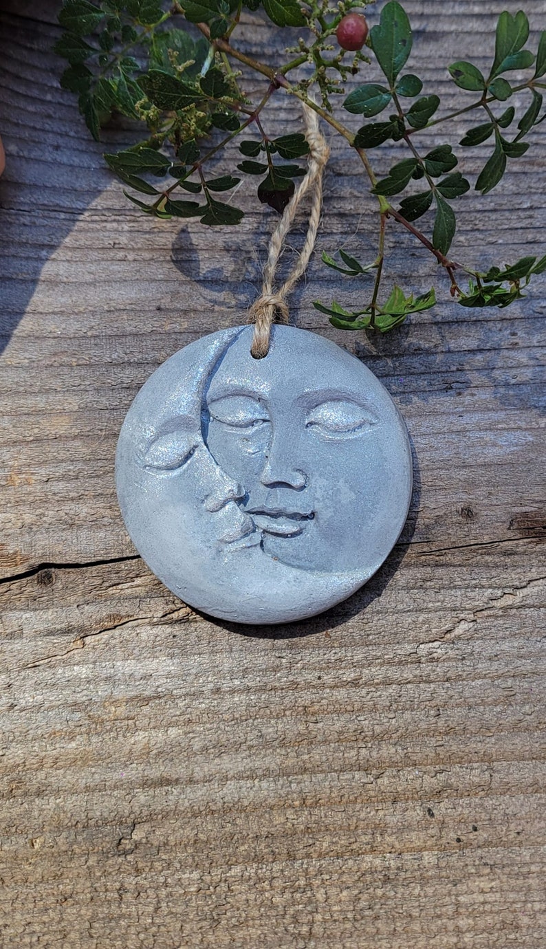 May include: A gray ceramic ornament shaped like a crescent moon with a face on it. The moon is hanging from a twine string.