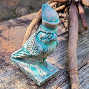 May include: A teal falcon statue, possibly depicting the Egyptian god Horus. The bird is on a rectangular base with hieroglyphs. The statue has a weathered appearance, with a textured surface. The statue is made of a teal-colored material.