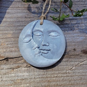 May include: A gray ceramic ornament shaped like a crescent moon with a face on it. The moon is hanging from a twine string.