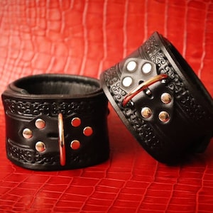 May include: A pair of black leather cuffs featuring an intricate embossed pattern, silver-toned metal rivets, and a central D-ring closure. The cuffs are constructed with a smooth interior lining and a textured exterior band design.