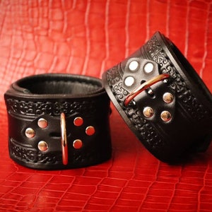 May include: Two black leather cuffs with silver buckles and decorative stitching. The cuffs are on a red background.