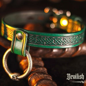 K&ouml;nnte beinhalten: Ein gr&uuml;nes Lederhalsband mit einem keltischen Knotendesign und einem silbernen Schlangenring. Das Halsband liegt auf einer Holzoberfl&auml;che. "Devilish Devices" ist in gotischer Schrift unter dem Halsband geschrieben.
