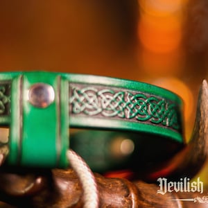 K&ouml;nnte beinhalten: Ein gr&uuml;nes Lederhalsband mit einem keltischen Knotendesign und einer silbernen Metallschnalle. Das Halsband liegt auf einer Holzoberfl&auml;che.