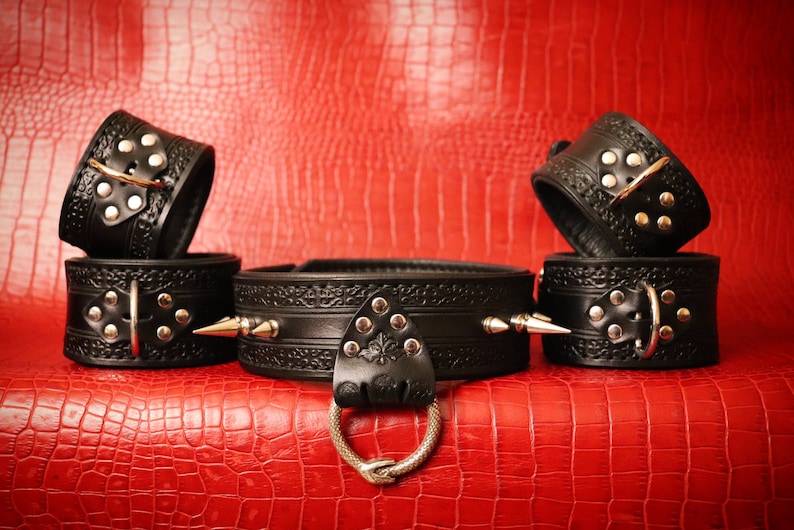 May include: Black leather collar and cuffs with silver hardware and studs. The collar has a decorative ring on the front.