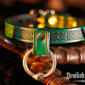 K&ouml;nnte beinhalten: Ein gr&uuml;nes Lederhalsband mit einem keltischen Knotendesign und einem silbernen Ring. Das Halsband liegt auf einer Holzoberfl&auml;che. "Devilish Devices" ist in gotischer Schrift unter dem Halsband geschrieben.