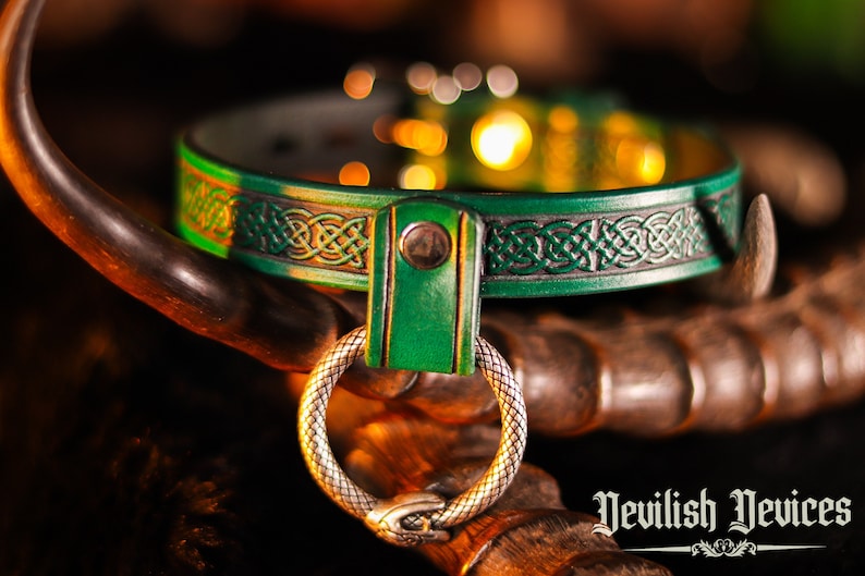 K&ouml;nnte beinhalten: Ein gr&uuml;nes Lederhalsband mit einem keltischen Knotendesign und einem silbernen Ring. Das Halsband liegt auf einer Holzoberfl&auml;che. "Devilish Devices" ist in gotischer Schrift unter dem Halsband geschrieben.