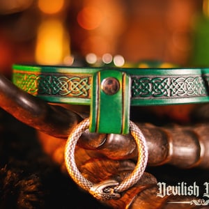 K&ouml;nnte beinhalten: Ein gr&uuml;nes Lederhalsband mit einem keltischen Knotendesign und einem silbernen Ring. Das Halsband liegt auf einer braunen Oberfl&auml;che mit dem Text "Nevilish Devices" unten rechts.