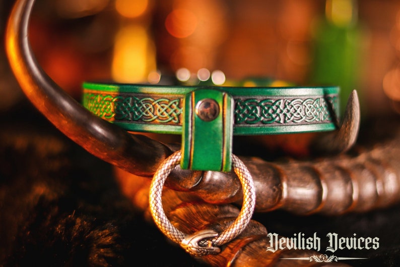 K&ouml;nnte beinhalten: Ein gr&uuml;nes Lederhalsband mit einem keltischen Knotendesign und einem silbernen Ring. Das Halsband liegt auf einer braunen Oberfl&auml;che mit dem Text "Nevilish Devices" unten rechts.