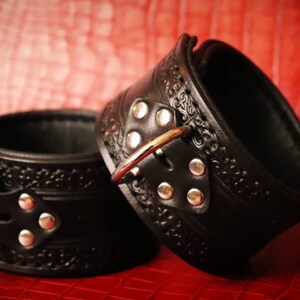 May include: Two black leather cuffs with silver buckles and decorative embossed designs. The cuffs are on a red surface.