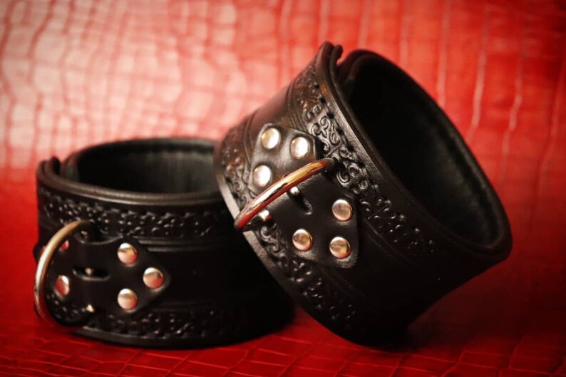 May include: Two black leather cuffs with silver buckles and decorative embossed designs. The cuffs are on a red surface.