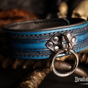 May include: A blue leather collar with a silver ring and black accents. The collar has a decorative pattern and is made of high-quality leather.