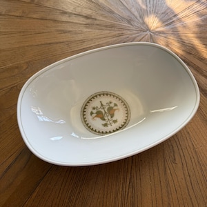May include: White oval serving bowl with a decorative floral design in the center. The bowl has a thin green trim around the rim and a detailed pattern in the center with green, orange, and brown elements. The bowl is set on a wooden surface.