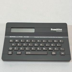 May include: A dark gray Franklin Computer Spelling Ace electronic spelling device. It features a QWERTY keyboard layout, a small display screen, and the text "SPELLING ACE" and "Linguistic Technology by Proximity/Merriam-Webster".