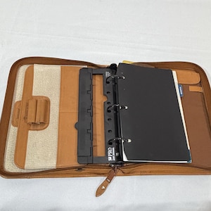 May include: A tan zippered portfolio organizer featuring an interior three-ring binder mechanism with a black plastic spine labeled PRO. The interior includes a textured cream-colored panel, a pen loop, and a black cover sheet for the binder.