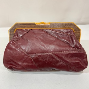 May include: A burgundy leather clutch bag with a geometric design. The bag has a wooden frame with a yellow clasp. The leather is stitched together in a patchwork style. The bag is closed.