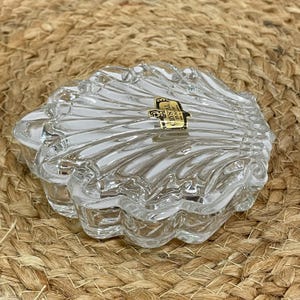 May include: A clear, crystal seashell-shaped trinket box. The box has a lid and is made of clear glass with a textured, ridged design. A gold and black label is affixed to the top. The box is sitting on a woven, tan surface.