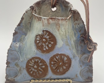 May include: Handmade ceramic vase with an irregular shape. The vase has a light blue and brown glaze with three brown floral designs. A leather strap is attached for hanging. The vase is displayed on a gold metal stand.