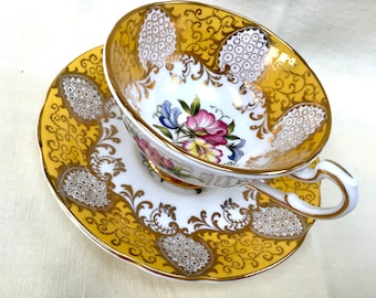 May include: A white teacup and saucer with a gold and yellow floral pattern. The cup and saucer have a delicate lace design around the rim.