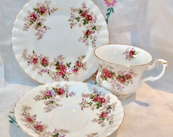 May include: A white porcelain tea set consisting of a teacup, a saucer, and a small plate. Each piece features a scalloped edge, gold trim, and a delicate pattern of pink roses with green leaves and brown fern-like accents on a white background.