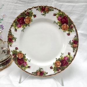 Puede incluir: Un plato de porcelana blanca con un borde dorado y un diseño floral con rosas rojas, amarillas y verdes. El plato está sobre un soporte blanco.