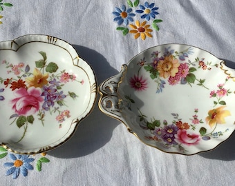 May include: Two white porcelain serving dishes with floral patterns in shades of pink, yellow, and purple. The dishes have gold trim and scalloped edges. One dish has two handles.