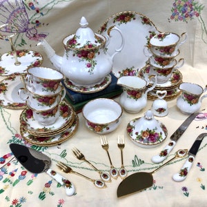 Puede incluir: Un juego de té vintage con un patrón floral, que incluye una tetera, una jarra de crema, un azucarero, tazas, platillos y un soporte para pasteles. El juego está sobre un mantel blanco con un patrón floral.