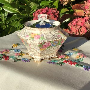 May include: A vintage, cream-colored porcelain trinket box with a floral pattern and gold trim. The box has a faceted shape and a clear, crystal-like knob on the lid. The box is sitting on a white tablecloth with colorful embroidery.