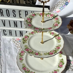 May include: A three-tier serving stand featuring white porcelain plates with scalloped edges and a floral pattern of pink roses and green leaves. The stand has a central gold-coloured metal rod and a decorative handle at the top for carrying.