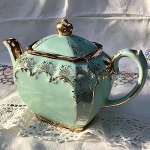 May include: A vintage, pale-blue teapot with gold trim and floral detailing. The teapot has a gold crown on the lid and a curved handle. The design includes gold detailing around the edges and on the handle.