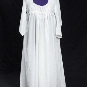 May include: A white, long-sleeved dress with a square neckline and gathered bodice. The dress is displayed on a purple dress form against a black backdrop. The sleeves are long and end in a slight flare.