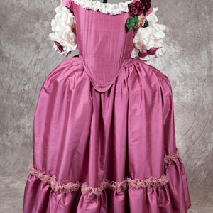 May include: A pink silk dress with a white lace trim and a floral embellishment on the shoulder. The dress has a fitted bodice and a full skirt with multiple layers of ruffles.