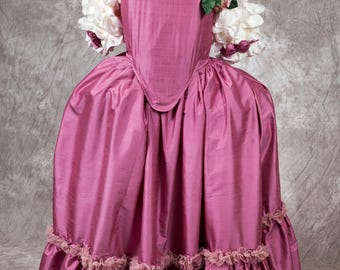 May include: A pink silk dress with a white lace trim and a floral embellishment on the shoulder. The dress has a fitted bodice and a full skirt with multiple layers of ruffles.