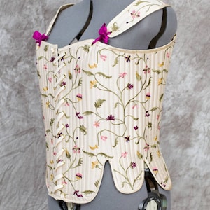 May include: A white corset with a floral pattern and pink ribbon accents. The corset is on a gray dress form.