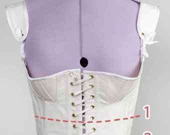 Puede incluir: Un corsé blanco con tirantes y espalda con cordones se muestra en un maniquí morado. El corsé presenta ojales dorados y un diseño estructurado. Las correas están aseguradas con pequeños lazos.