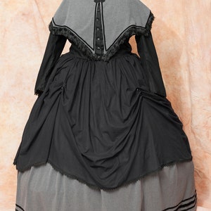 May include: A black and gray Victorian-style dress with a cape. The dress has a black skirt with a gray underskirt and a gray cape with black trim.