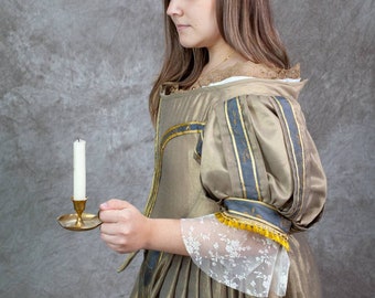May include: A woman in a gold and blue gown holds a brass candlestick with a white candle. The gown has a lace trim and a gathered skirt.
