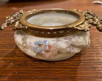 May include: A small, white porcelain trinket box with a gold-toned metal lid and floral design. The lid has a decorative, ornate design.