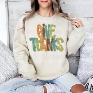 May include: Cream-colored sweatshirt with the words "GIVE THANKS" in colorful, layered lettering. The text features orange, green, and brown hues. The sweatshirt is a casual, long-sleeved garment, perfect for fall or Thanksgiving.
