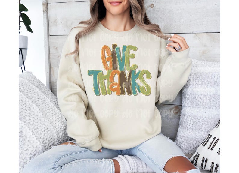 May include: Cream-colored sweatshirt with the words "GIVE THANKS" in colorful, layered lettering. The text features orange, green, and brown hues. The sweatshirt is a casual, long-sleeved garment, perfect for fall or Thanksgiving.