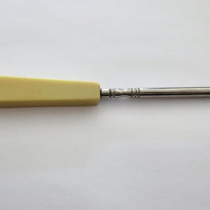 Puede incluir: Instrumento quirúrgico antiguo de color marfil con un gancho y mango de metal. El mango tiene forma rectangular y una pequeña inscripción. El gancho de metal está en el extremo de un eje metálico largo y delgado.