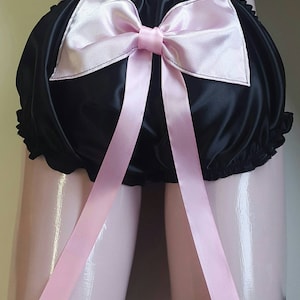 May include: Black satin underwear with a large pink bow and long pink ribbons. The underwear has a ruffled edge and is designed with a full coverage style. The bow is centered on the front, adding a decorative touch.
